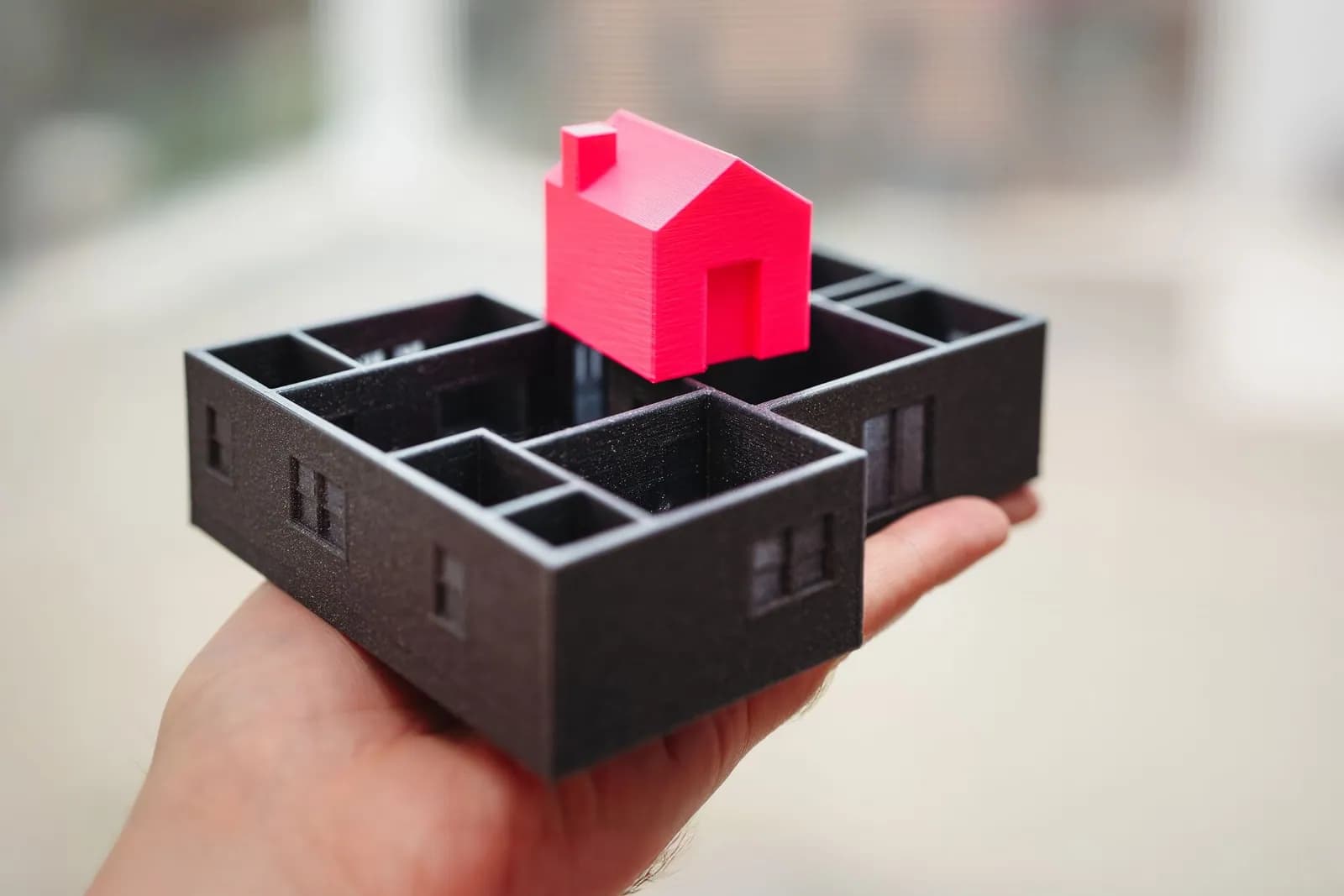 Hand hält ein 3D-gedrucktes Hausmodell über einem ausgedruckten Grundriss