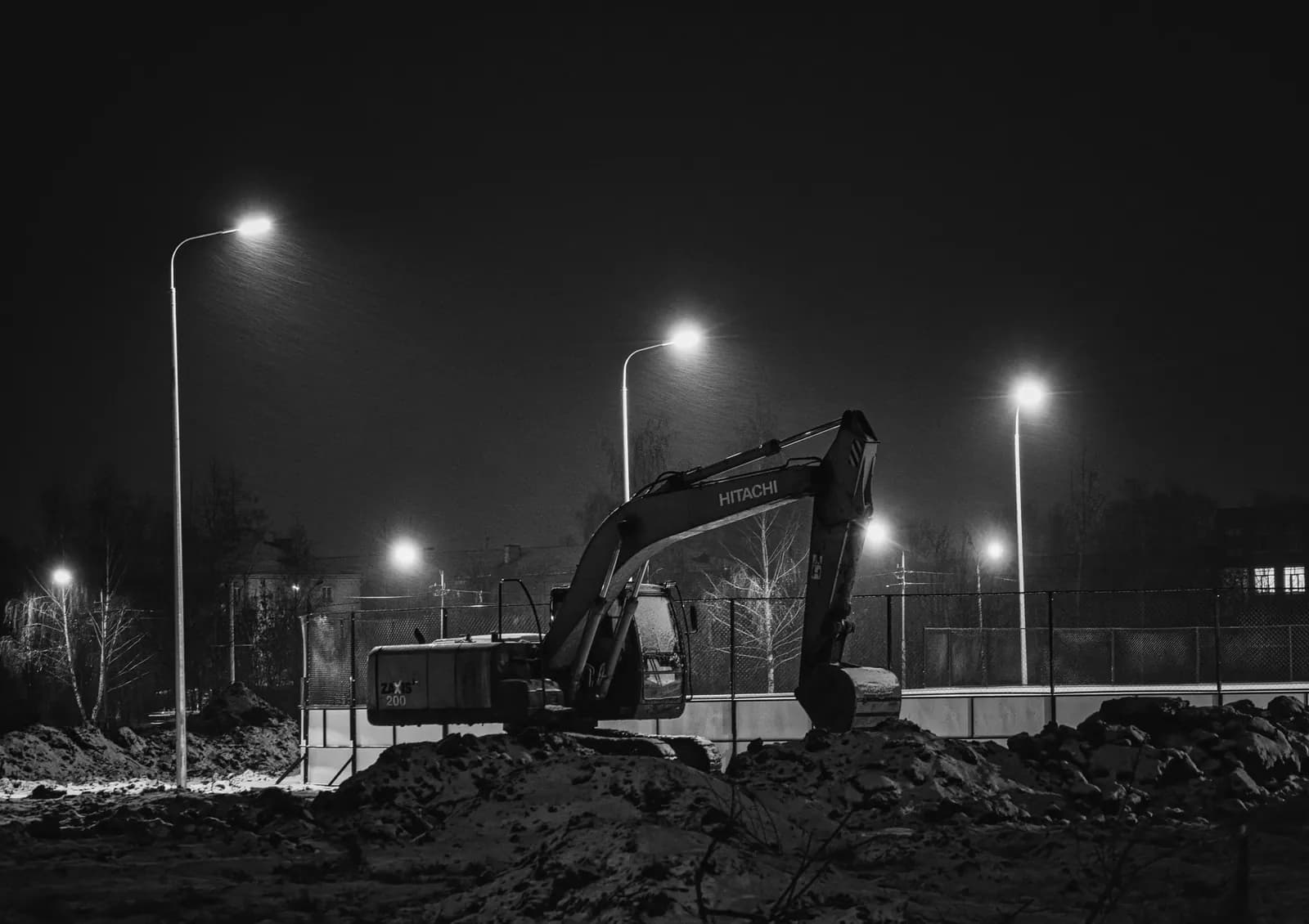 Bagger auf einer unbeleuchteten Freifläche bei Nacht unter Straßenlaternen, Schwarz-Weiß-Aufnahme