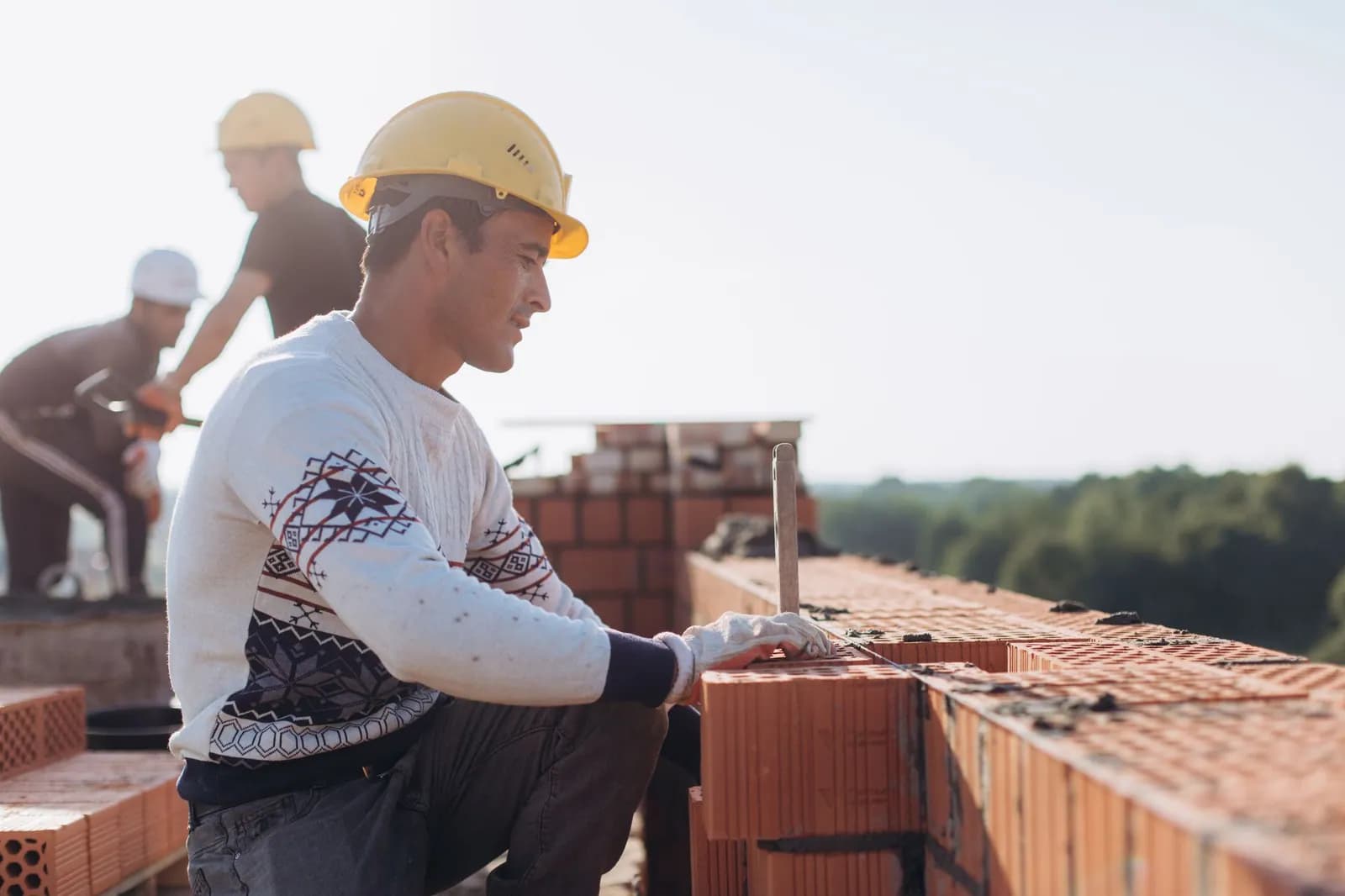 Bauarbeiter mauert im Freien eine Ziegelwand auf einer sonnigen Baustelle mit Grün im Hintergrund