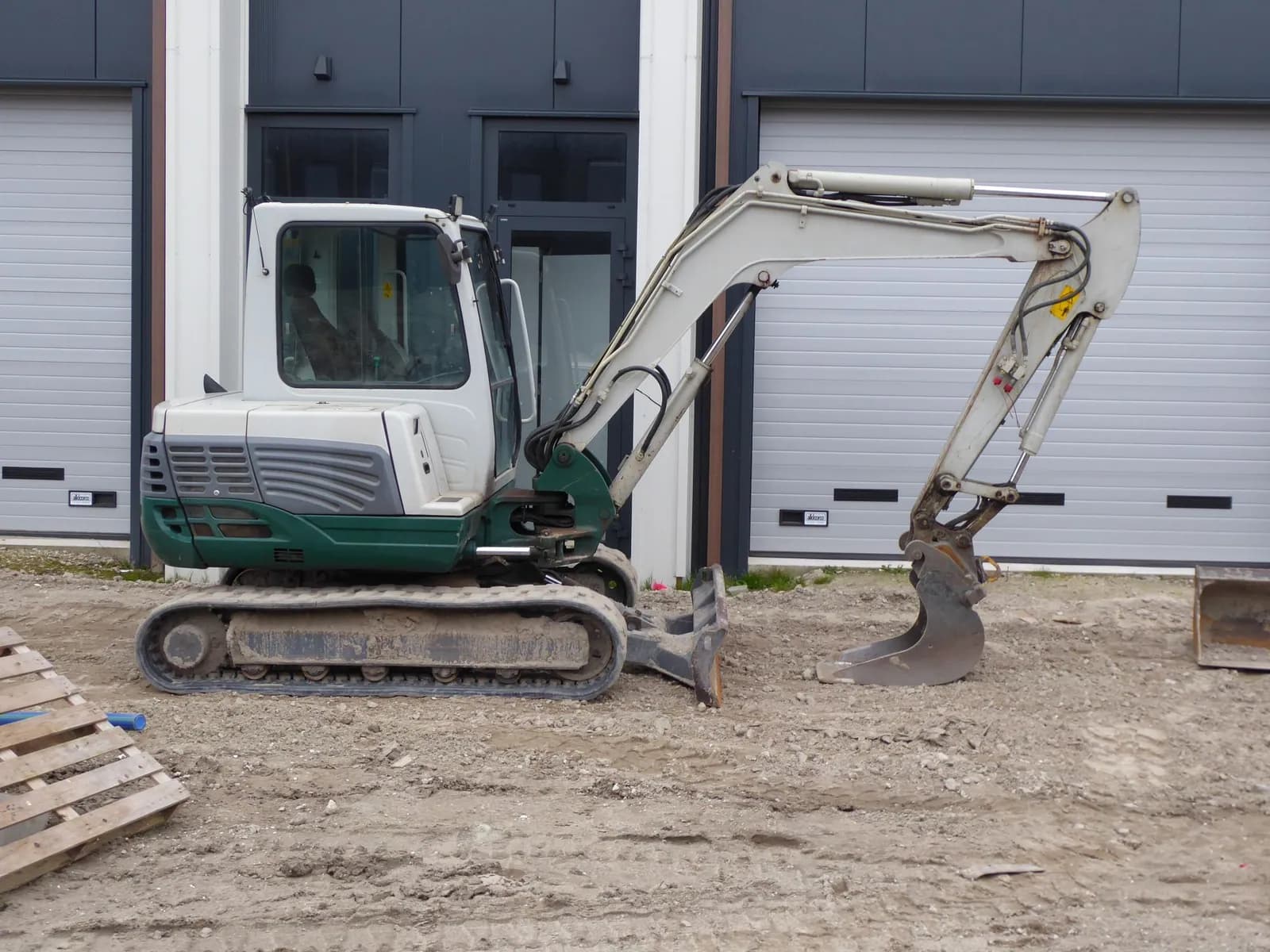 Kompaktbagger mit Raupenfahrwerk steht auf einer Baustelle vor einem Gebäude mit Rolltoren