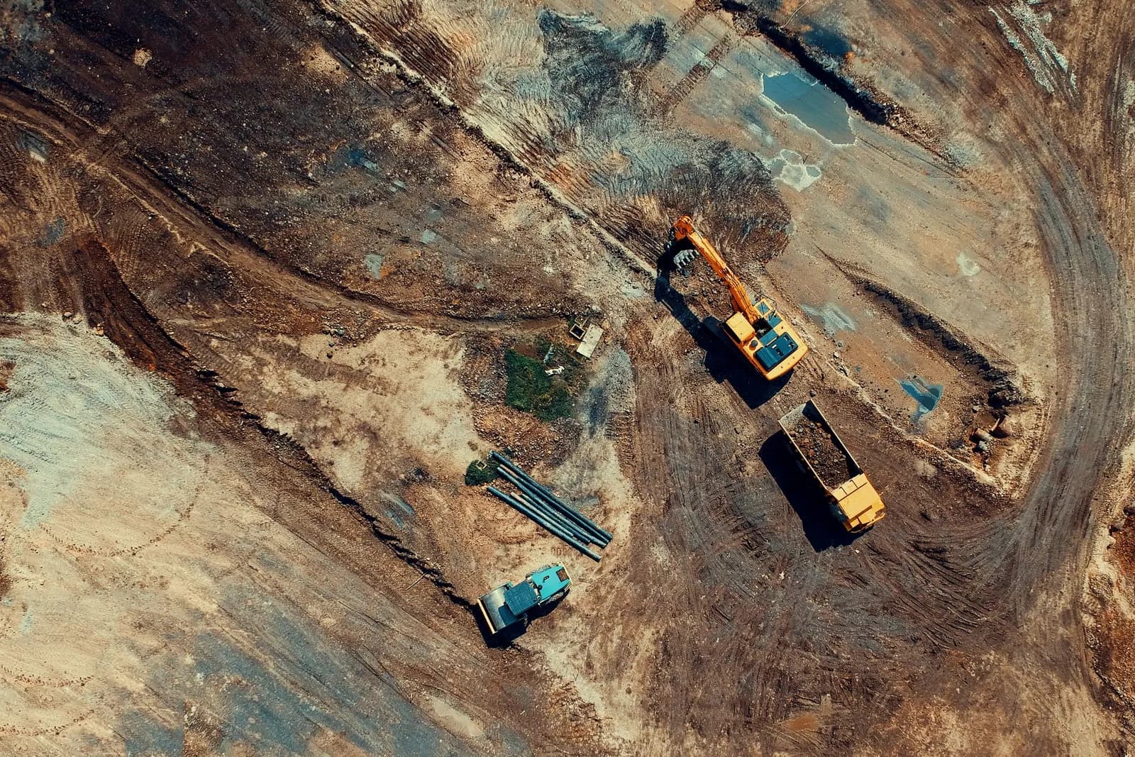 Luftaufnahme einer Tiefbaustelle: Bagger belädt einen Muldenkipper, daneben Walze und gelagerte Rohre